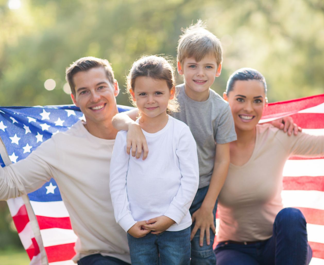 A family outdoors smiling with two adults and two children, holding an American flag behind them, conveying a sense of patriotic pride and togetherness.